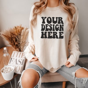 Pode incluir: Uma mulher vestindo uma camisa de manga comprida de cor creme com o texto "YOUR DESIGN HERE" em preto. Ela está vestindo jeans azul com rasgos nos joelhos.