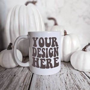 Puede incluir: Taza de cerámica blanca con un diseño negro que dice "YOUR DESIGN HERE" sobre una superficie de madera blanca con calabazas blancas.