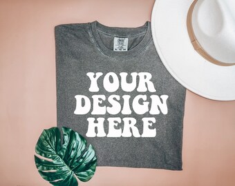 May include: A gray t-shirt with the text "YOUR DESIGN HERE" in white. The shirt is folded and laid flat on a pink surface. A white hat and a green leaf are also in the image.