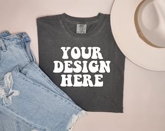May include: A dark gray t-shirt with the text "YOUR DESIGN HERE" in white, retro-style font. The shirt is laid flat on a light pink surface.