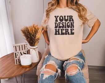 May include: A beige t-shirt with the text "YOUR DESIGN HERE" in black retro-style lettering. The shirt is being worn by a person with long blonde hair and blue jeans with distressed details.