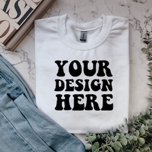 May include: White t-shirt with the text "YOUR DESIGN HERE" in black retro-style font. The shirt is laid flat on a marble surface with a pair of blue jeans and a sprig of greenery.