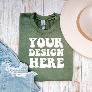 May include: A green t-shirt with the text "YOUR DESIGN HERE" in white. The shirt is folded and laid on a white wooden surface with a pair of blue jeans and a white hat.