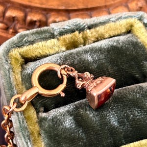 May include: A gold-colored antique pendant with a small, rectangular, reddish-brown stone. The pendant is attached to a gold chain and a clasp. The pendant is set against a green velvet background.