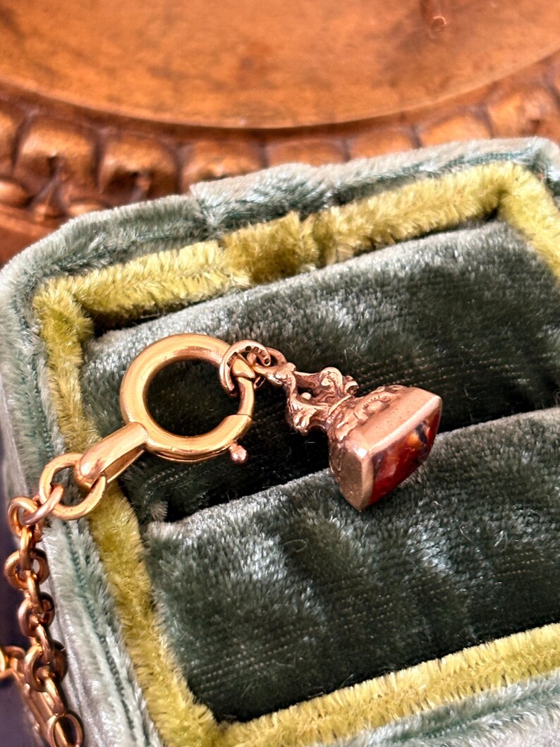 May include: A gold-colored antique pendant with a small, rectangular, reddish-brown stone. The pendant is attached to a gold chain and a clasp. The pendant is set against a green velvet background.