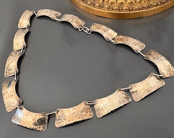 May include: A silver-toned necklace with a hammered texture. The necklace features rectangular, curved links connected by small metal rings. The jewelry is displayed on a dark surface, with a gold-colored decorative object in the background.