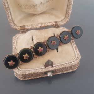 Puede incluir: Un broche vintage de ónix negro y oro con detalles de estrellas rojas. El broche está en una pequeña caja de terciopelo marrón.