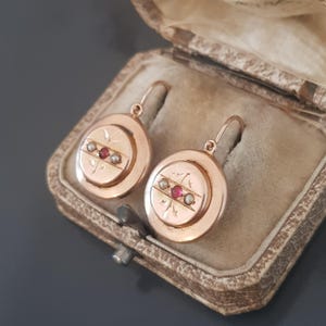 May include: A pair of gold drop earrings with a circular design. The earrings feature three red gemstones and two white pearls set in a gold band.
