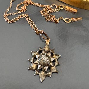 May include: A rose gold chain necklace featuring a star-shaped pendant with a textured, dark metallic finish. The pendant has a central raised point surrounded by multiple radiating spikes, and the chain includes a rectangular clasp component.