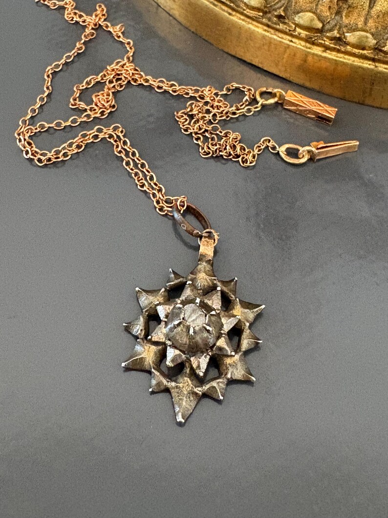 May include: A rose gold chain necklace featuring a star-shaped pendant with a textured, dark metallic finish. The pendant has a central raised point surrounded by multiple radiating spikes, and the chain includes a rectangular clasp component.