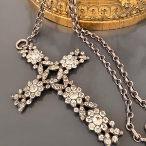 May include: A silver-toned necklace featuring a cross pendant adorned with numerous clear, sparkling crystals. The cross design incorporates floral-like clusters of crystals, and the chain is a delicate link style. The necklace rests on a gray surface.