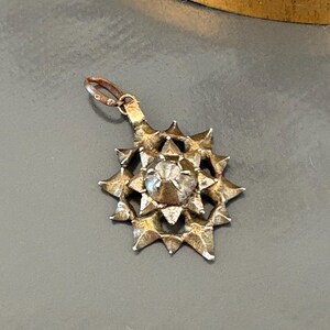 May include: A silver-toned metal pendant shaped like a multi-pointed star with a textured, jagged surface. The center features a raised, faceted, clear stone setting. A small, circular jump ring is attached to the top for hanging purposes.