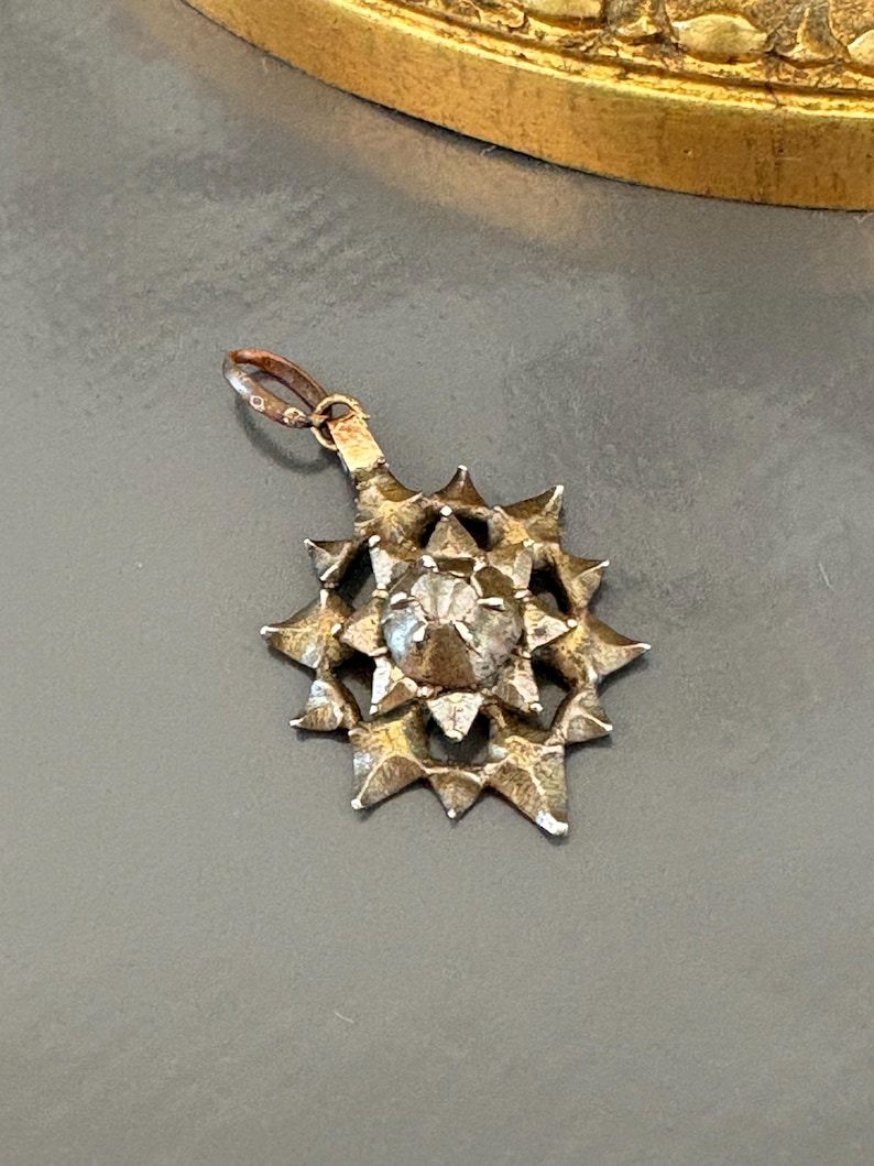 May include: A silver-toned metal pendant shaped like a multi-pointed star with a textured, jagged surface. The center features a raised, faceted, clear stone setting. A small, circular jump ring is attached to the top for hanging purposes.