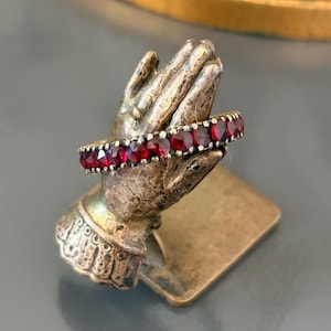 Op de afbeelding: Een ring in vintage-stijl met een band van kleine, ronde, dieprode edelstenen. De ring wordt getoond op een kleine, antiek ogende handvormige standaard. De standaard is gemaakt van een bronskleurig metaal en heeft ingewikkelde details.