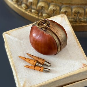 May include: A round, reddish-brown nut-like object encased in a brass band with a small metal loop at the top, accompanied by two small, pointed, light-colored wooden pegs with metal bases, all resting on a square, off-white textured surface.