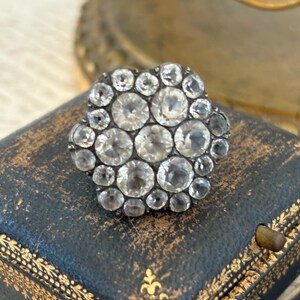 May include: A cluster ring featuring numerous round, clear, faceted stones arranged in a floral-like pattern. The stones are set in a dark, oxidized metal frame, creating a high-contrast look with the sparkling, transparent gems against the dark setting.