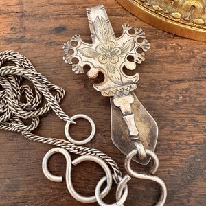 Puede incluir: Un colgante de metal plateado con una forma ornamentada similar a una cruz, con detalles florales grabados y bordes festoneados. El colgante está unido a una cadena plateada texturizada con múltiples eslabones circulares abiertos y conectores en forma de S.