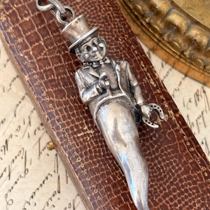 May include: A silver-toned charm in the shape of a gentleman wearing a top hat and suit, holding a small object and a horseshoe. The charm is attached to a chain with a clasp. The charm is on a brown leather surface.