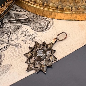 May include: A silver-toned metal pendant featuring a multi-pointed star design with a central circular setting holding a single stone. The pendant includes a small circular jump ring at the top for attachment, showcasing a textured, dark metallic finish.