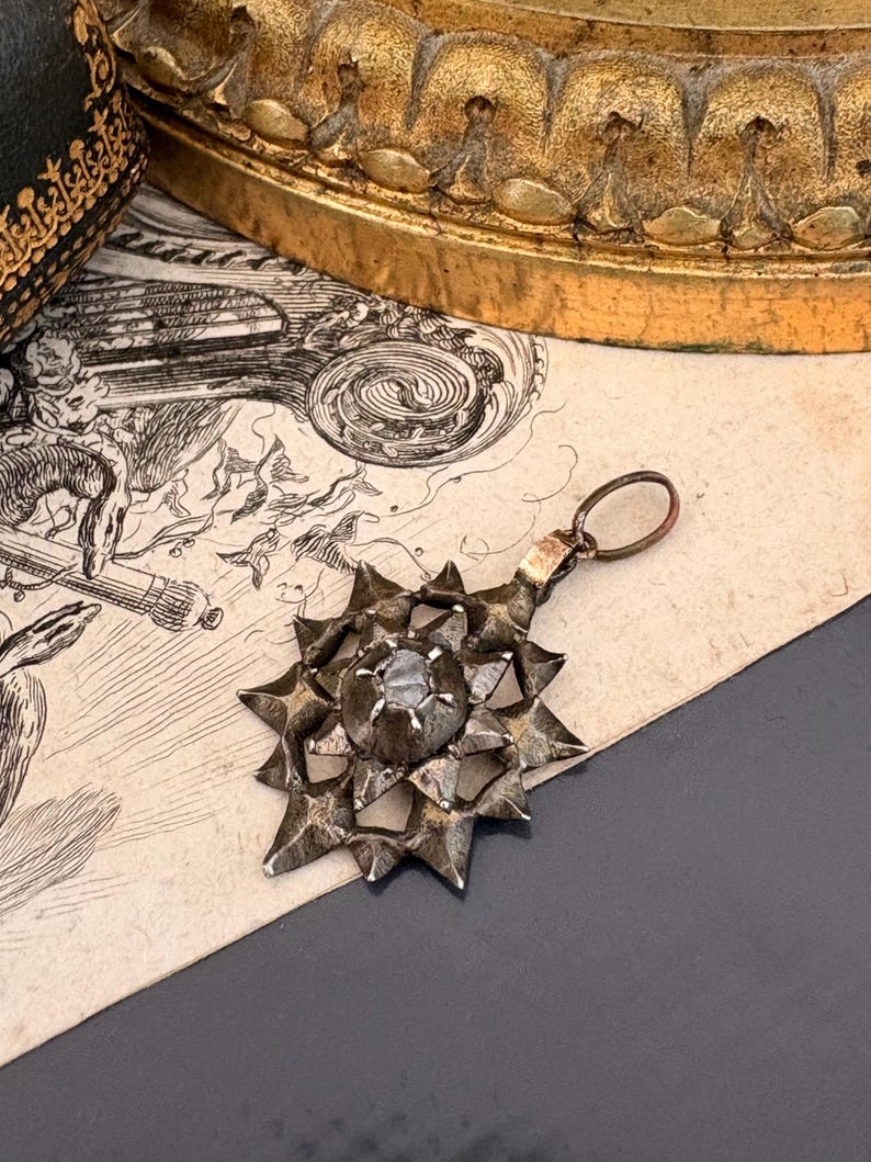 May include: A silver-toned metal pendant featuring a multi-pointed star design with a central circular setting holding a single stone. The pendant includes a small circular jump ring at the top for attachment, showcasing a textured, dark metallic finish.