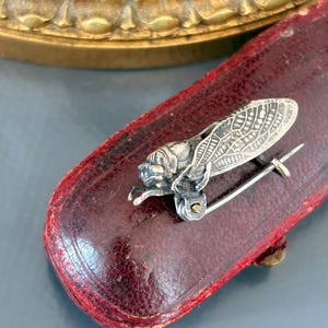 Puede incluir: Broche de plata con forma de cigarra, con intrincados detalles en las alas, sobre un estuche de cuero burdeos. El alfiler de insecto tiene un cierre y un alfiler para una fijación segura. Un accesorio vintage para un toque de elegancia.