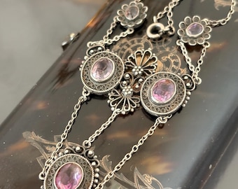 Puede incluir: Un collar plateado con piedras preciosas de amatista ovaladas y en forma de flor. El collar presenta intrincados detalles de filigrana alrededor de las piedras preciosas y delicadas cadenas. La joya se exhibe sobre una superficie oscura y estampada.