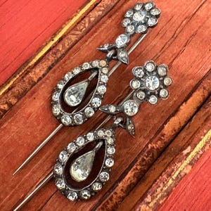 May include: Two antique silver brooches. Each brooch features a teardrop-shaped pendant with a central clear stone, surrounded by smaller clear stones. The top of each brooch has a floral design with more clear stones. The brooches are set against a reddish-brown background.