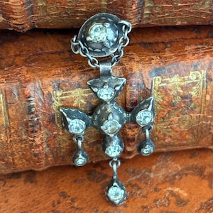 Può includere: Ciondolo antico color argento con pietre preziose rotonde e trasparenti. Il ciondolo presenta una parte superiore circolare collegata da una catena a un design a forma di croce con gemme a goccia. Su uno sfondo di vecchi libri rilegati in pelle.