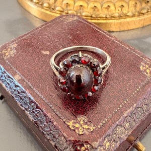 Può includere: Un anello in argento con una grande pietra centrale color rosso scuro a cabochon, circondata da un alone circolare di pietre più piccole, sfaccettate e di colore rosso intenso. L'anello presenta una sottile fascia in argento lucido.