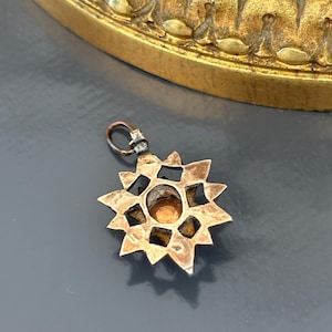 May include: A star-shaped metal pendant featuring a central circular amber-colored stone. The pendant has a textured, aged metallic finish with a series of triangular points radiating from the center and includes a small jump ring at the top.