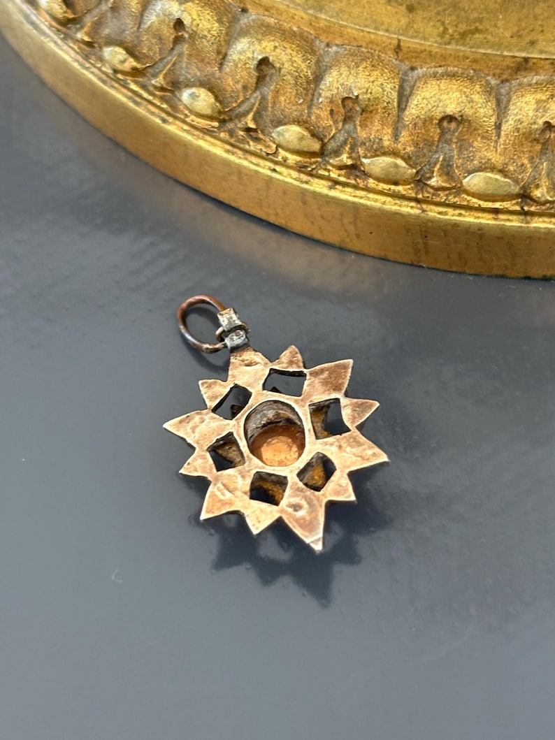May include: A star-shaped metal pendant featuring a central circular amber-colored stone. The pendant has a textured, aged metallic finish with a series of triangular points radiating from the center and includes a small jump ring at the top.