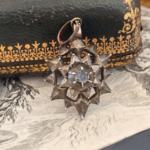 May include: A silver-toned metal pendant shaped like a multi-pointed star with a central circular setting containing a single light-colored stone. The pendant features a small circular bail at the top for attachment to a chain or cord.
