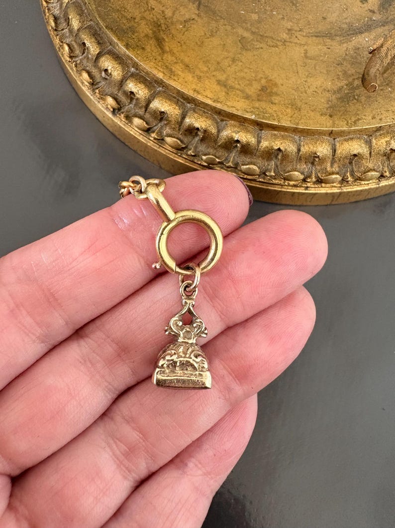 May include: A gold-colored antique fob seal with a detailed, ornate design. The seal is suspended from a circular clasp and chain. The background features a blurred, decorative gold-toned object.