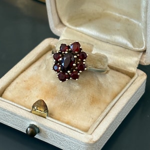 May include: A vintage gold ring with a cluster of deep red gemstones, displayed in a cream velvet-lined jewellery box. The ring features a central, larger stone surrounded by smaller gems. The box is marked "MELUN".