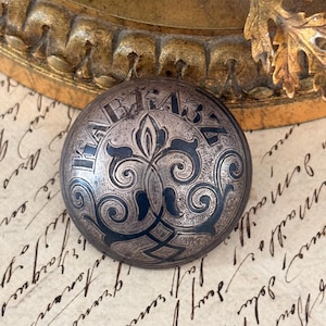 May include: A circular, metallic button with a domed surface featuring an intricate, dark-toned engraved floral pattern and the Cyrillic text "КАВКАЗЪ" arched across the top. The metal has a weathered, antique finish with visible oxidation.