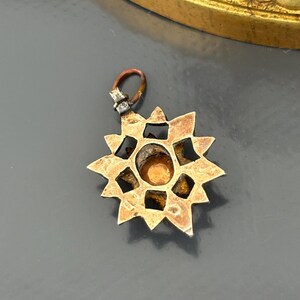 May include: A star-shaped metal pendant featuring a central circular amber-colored stone set within a geometric frame. The pendant has a weathered, metallic finish and includes a small, dark-toned jump ring attached to the top for suspension.