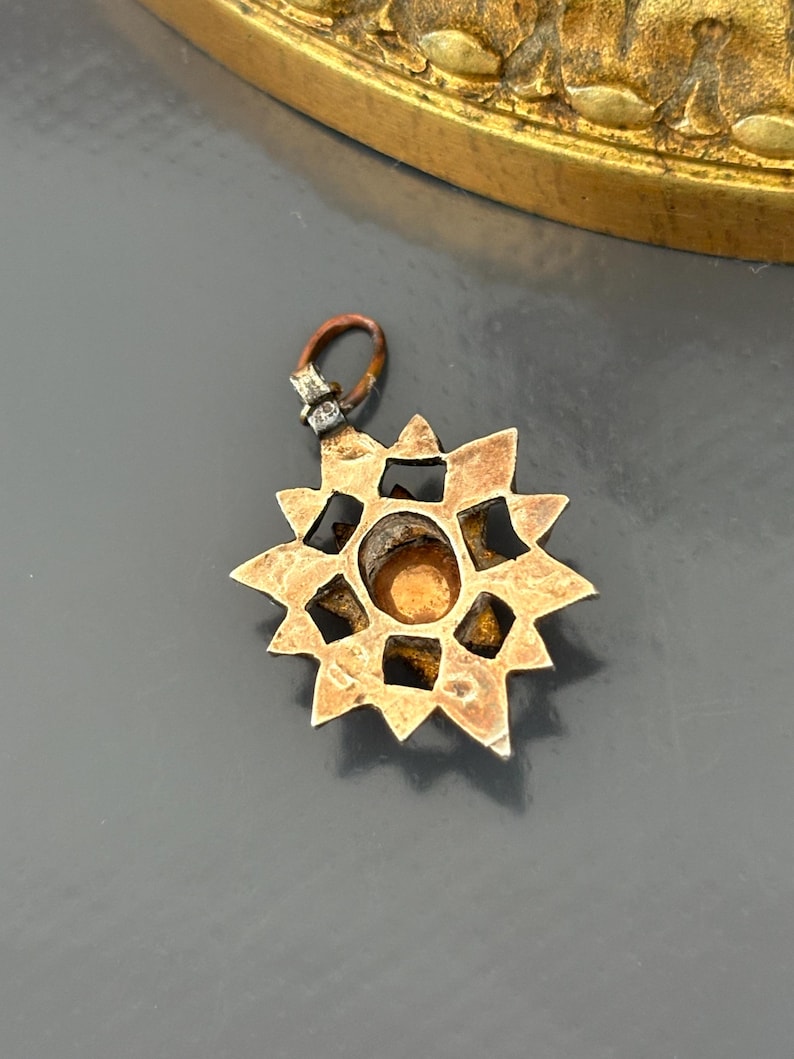 May include: A star-shaped metal pendant featuring a central circular amber-colored stone set within a geometric frame. The pendant has a weathered, metallic finish and includes a small, dark-toned jump ring attached to the top for suspension.