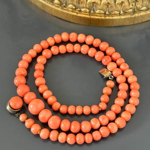 May include: A coral bead necklace with a gold clasp. The necklace features three strands of round, peach-colored beads of varying sizes. The necklace is displayed on a gray surface.