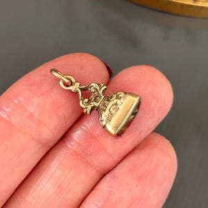 May include: A small, ornate gold-colored pendant with a detailed design. The pendant has a rectangular base and a decorative top with a loop for a chain. The pendant is held in a hand against a gray background.