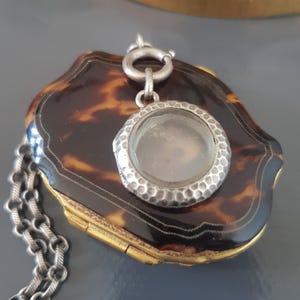 Puede incluir: Un medallón de plata con un exterior de caparazón de tortuga y una ventana de vidrio. El medallón está unido a una cadena de plata.
