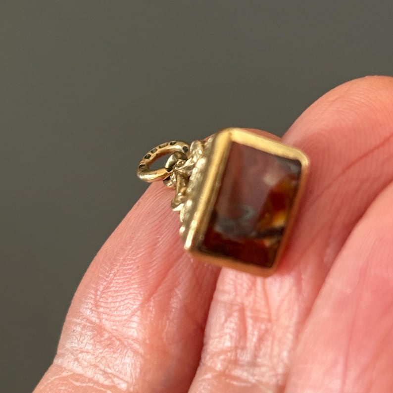 May include: A gold-colored pendant with a rectangular, reddish-brown stone. The pendant has intricate detailing around the stone and a small loop for a chain. The stone has a marbled appearance.
