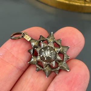 May include: A silver-toned metal pendant featuring a multi-pointed star design with a raised, textured spherical center. The pendant includes a small circular bail at the top for attachment, showcasing a weathered, metallic finish throughout the piece.
