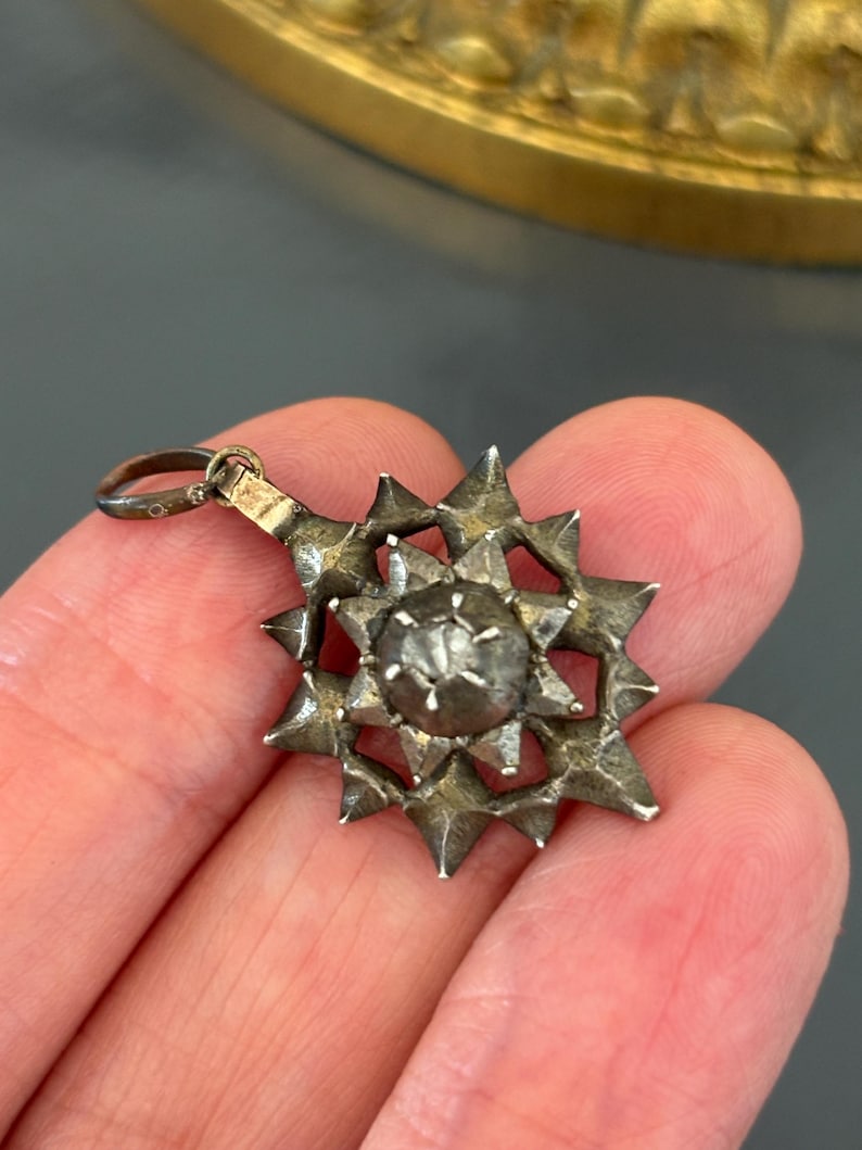 May include: A silver-toned metal pendant featuring a multi-pointed star design with a raised, textured spherical center. The pendant includes a small circular bail at the top for attachment, showcasing a weathered, metallic finish throughout the piece.