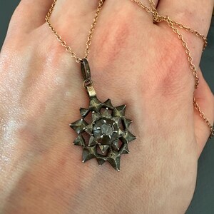May include: A silver-toned star-shaped pendant with a central circular stone, suspended from a thin gold-colored metal chain. The pendant features a multi-pointed, textured design with a dark, weathered finish, hanging from a simple metal bail.