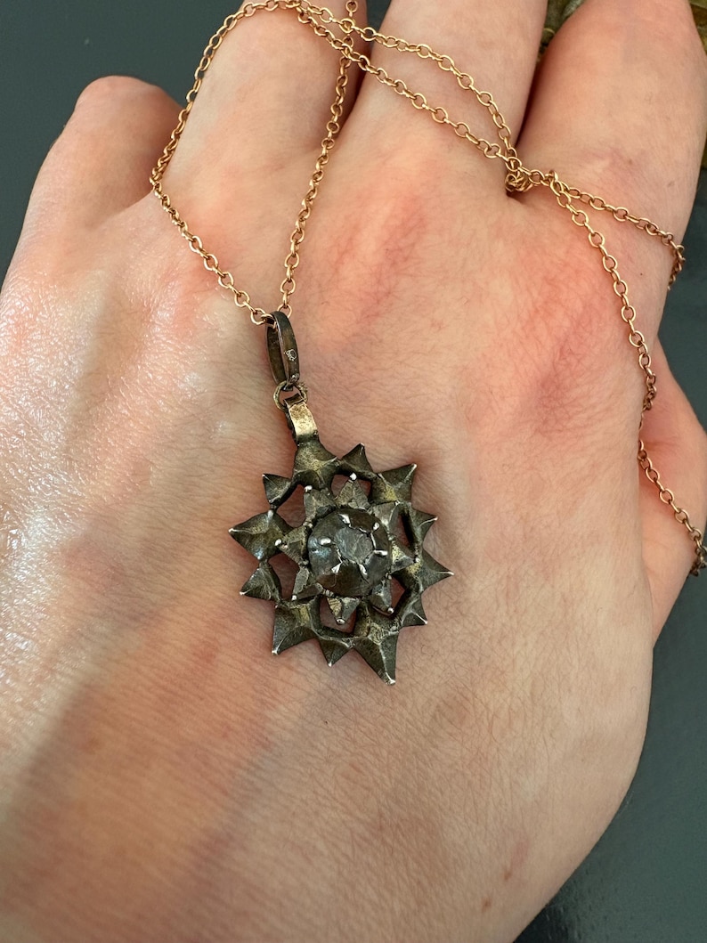May include: A silver-toned star-shaped pendant with a central circular stone, suspended from a thin gold-colored metal chain. The pendant features a multi-pointed, textured design with a dark, weathered finish, hanging from a simple metal bail.