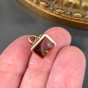 May include: A small, square pendant with a gold-colored frame and a reddish-brown stone. The pendant is held in a person's hand, with a gold loop at the top. The stone has a unique pattern of darker and lighter shades.