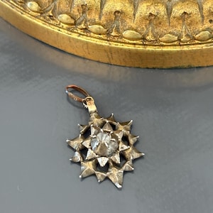 May include: A silver-toned metal pendant shaped like a multi-pointed star with a central faceted clear stone. The pendant features a textured, hammered surface and includes a small circular bail at the top for attachment to a chain or cord.