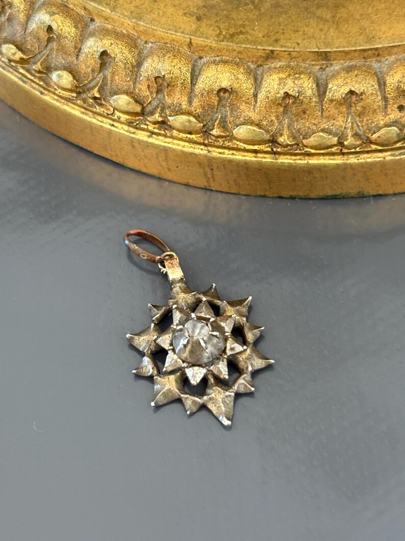 May include: A silver-toned metal pendant shaped like a multi-pointed star with a central faceted clear stone. The pendant features a textured, hammered surface and includes a small circular bail at the top for attachment to a chain or cord.