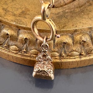 May include: Close-up of a gold-colored pendant with intricate scrollwork and a small, round clasp. The pendant hangs from a delicate chain, and the background features a textured, golden surface.