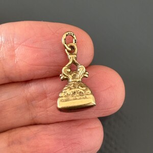 May include: A small, antique gold-colored pendant with a detailed design. The pendant has a loop at the top for a chain and a bell-shaped body with intricate carvings. The overall design is ornate and vintage.